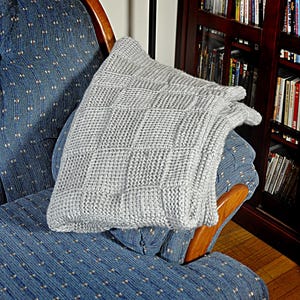 May include: A gray knitted throw blanket with a square pattern is draped over a blue patterned armchair. The armchair is made of wood and has a curved back.