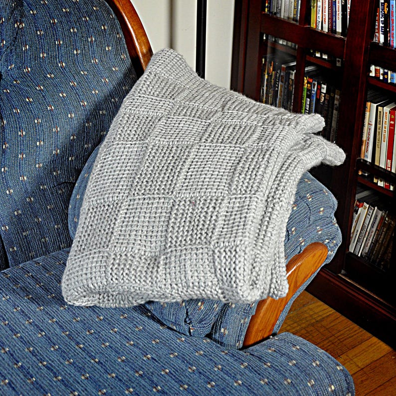 May include: A gray knitted throw blanket with a square pattern is draped over a blue patterned armchair. The armchair is made of wood and has a curved back.