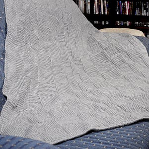 May include: A gray knitted throw blanket with a woven pattern. The blanket is draped over a chair.