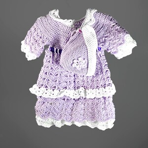 May include: A lavender crochet dress with white trim and a matching bonnet. The dress features a tiered skirt, short sleeves, and a purple ribbon at the waist. The bonnet is adorned with a small crochet flower detail on the side, completing the set.
