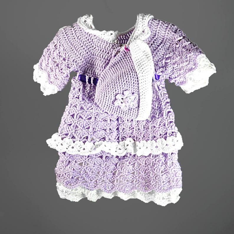 May include: A lavender crochet dress with white trim and a matching bonnet. The dress features a tiered skirt, short sleeves, and a purple ribbon at the waist. The bonnet is adorned with a small crochet flower detail on the side, completing the set.