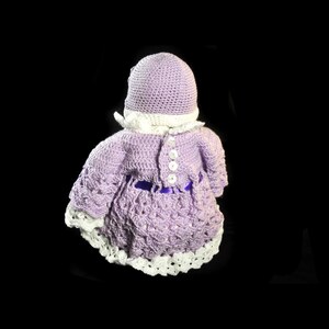 May include: A crochet doll wearing a lavender dress and matching bonnet. The dress features a white lace-trimmed hem, a purple ribbon waistband, and four white buttons down the back. The bonnet is also lavender with a white lace trim at the base.