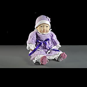 May include: A doll wearing a lavender crocheted dress with white lace trim, a matching lavender and white crocheted hat with a small flower detail, and a dark purple ribbon tied at the waist. The doll has light skin and wears gray socks.
