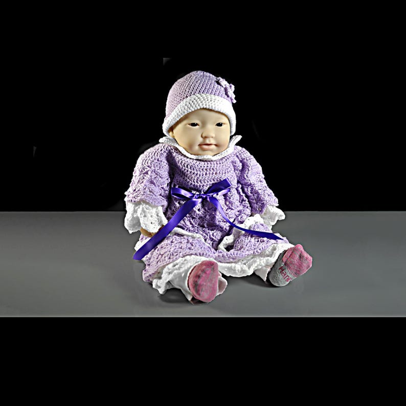 May include: A doll wearing a lavender crocheted dress with white lace trim, a matching lavender and white crocheted hat with a small flower detail, and a dark purple ribbon tied at the waist. The doll has light skin and wears gray socks.