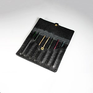 May include: A black leather case with a zipper closure, designed to hold and organize crochet hooks. The case is open, revealing six colorful crochet hooks inside.