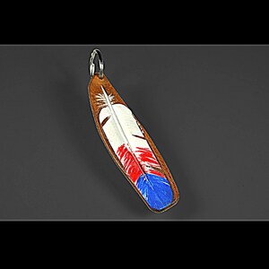 Puede incluir: Un llavero de cuero marrón con forma de pluma, pintado con detalles blancos, rojos y azules. Tiene un anillo de metal plateado para sujetar las llaves. Un accesorio decorativo.