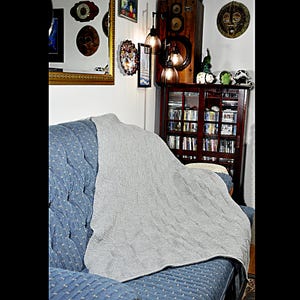 May include: A gray knitted throw blanket draped over a blue couch. The blanket has a cable knit pattern and is folded over the back of the couch.