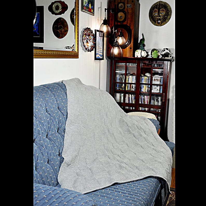 May include: A gray knitted throw blanket draped over a blue couch. The blanket has a cable knit pattern and is folded over the back of the couch.