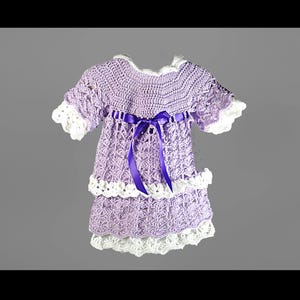 May include: A lavender crochet dress featuring a white scalloped trim on the sleeves and hem. The bodice includes a dark purple satin ribbon tied in a bow at the center. The skirt is constructed with two tiers of intricate crochet patterns.