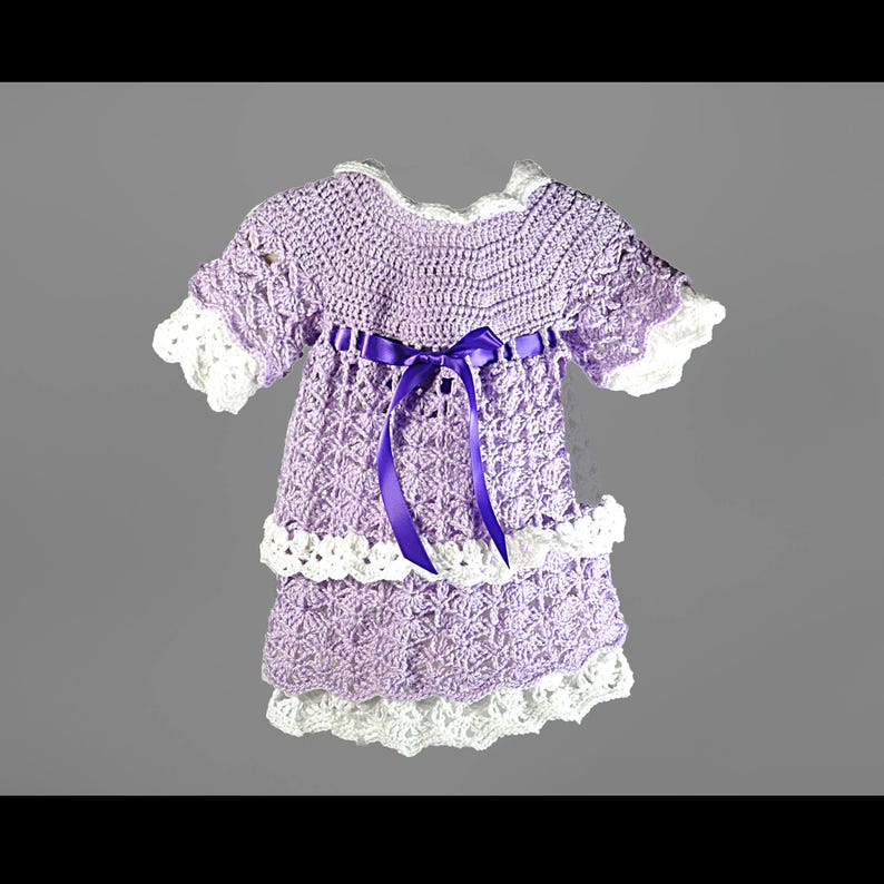 May include: A lavender crochet dress featuring a white scalloped trim on the sleeves and hem. The bodice includes a dark purple satin ribbon tied in a bow at the center. The skirt is constructed with two tiers of intricate crochet patterns.
