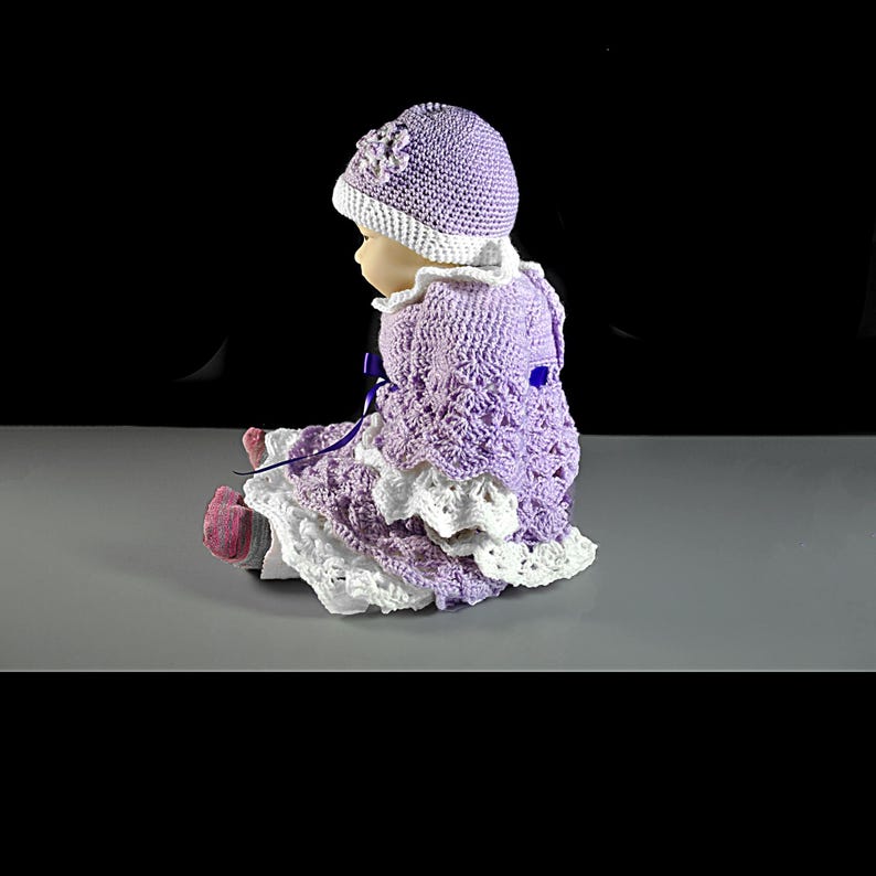 May include: A doll wearing a lavender and white crocheted outfit consisting of a matching hat with a flower detail, a long-sleeved dress with a textured pattern, and white lace-trimmed edges. The doll has dark pink socks and a purple ribbon tie.