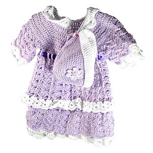 May include: A lavender and white crocheted dress featuring short sleeves, a white scalloped collar, and a white scalloped hem. A purple ribbon is threaded through the waist, and a matching lavender and white crocheted bonnet is included.