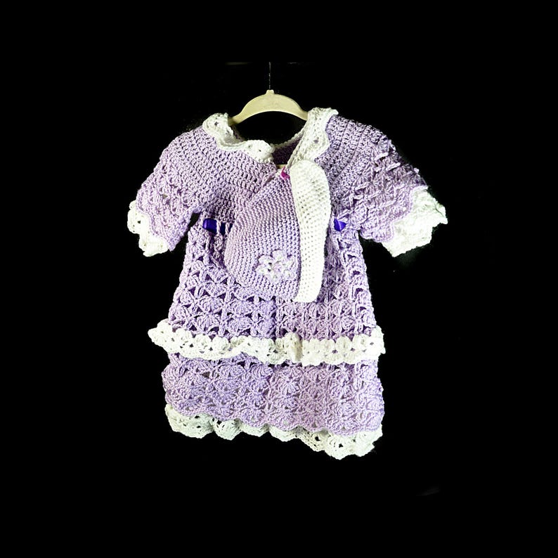 May include: A lavender crochet dress with short sleeves and a matching bonnet. The dress features white scalloped trim at the collar, sleeves, and hem, with a tiered skirt design and a small purple ribbon accent at the waist. The bonnet has a white trim.