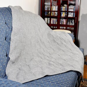 May include: A gray knitted throw blanket draped over a blue couch with a white polka dot pattern. The blanket has a woven texture and is folded over the back of the couch.