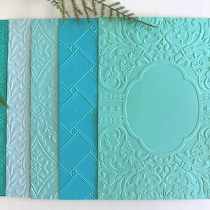 May include: A collection of embossed paper in shades of teal and blue. The papers feature various patterns, including floral, geometric, and ornate designs. A sprig of greenery is in the upper left corner.