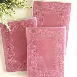 May include: Three pink decorative folders with intricate white floral and geometric border designs. The folders are arranged in a layered composition, with a sprig of white flowers in the upper left corner. The folders have a clear plastic overlay.