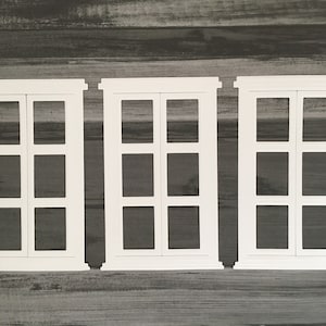 May include: Three white wooden window frames with a grid pattern of nine squares. The frames are set against a dark wood background.