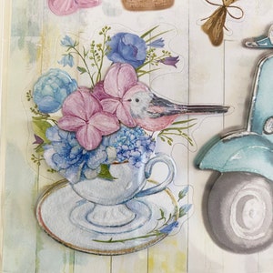 May include: A collection of floral-themed stickers featuring a teacup overflowing with blue and pink flowers, a bird, a hot air balloon basket, a bouquet, and a scooter. The stickers have a watercolor effect and are set against a light, textured background.