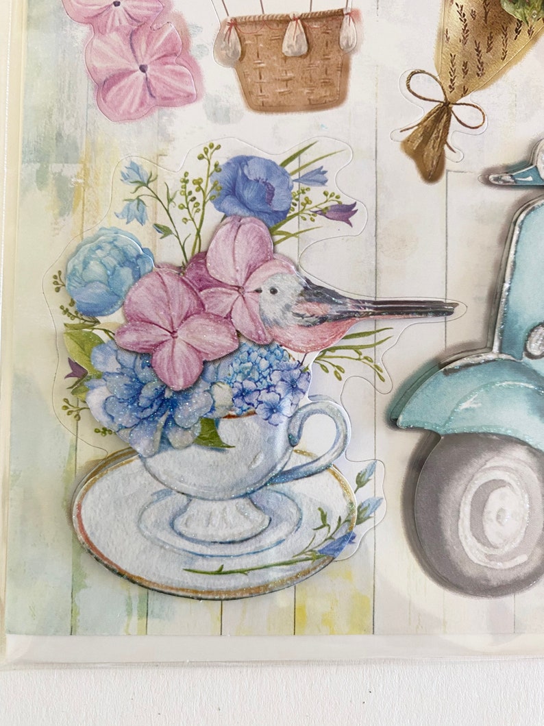 May include: A collection of floral-themed stickers featuring a teacup overflowing with blue and pink flowers, a bird, a hot air balloon basket, a bouquet, and a scooter. The stickers have a watercolor effect and are set against a light, textured background.