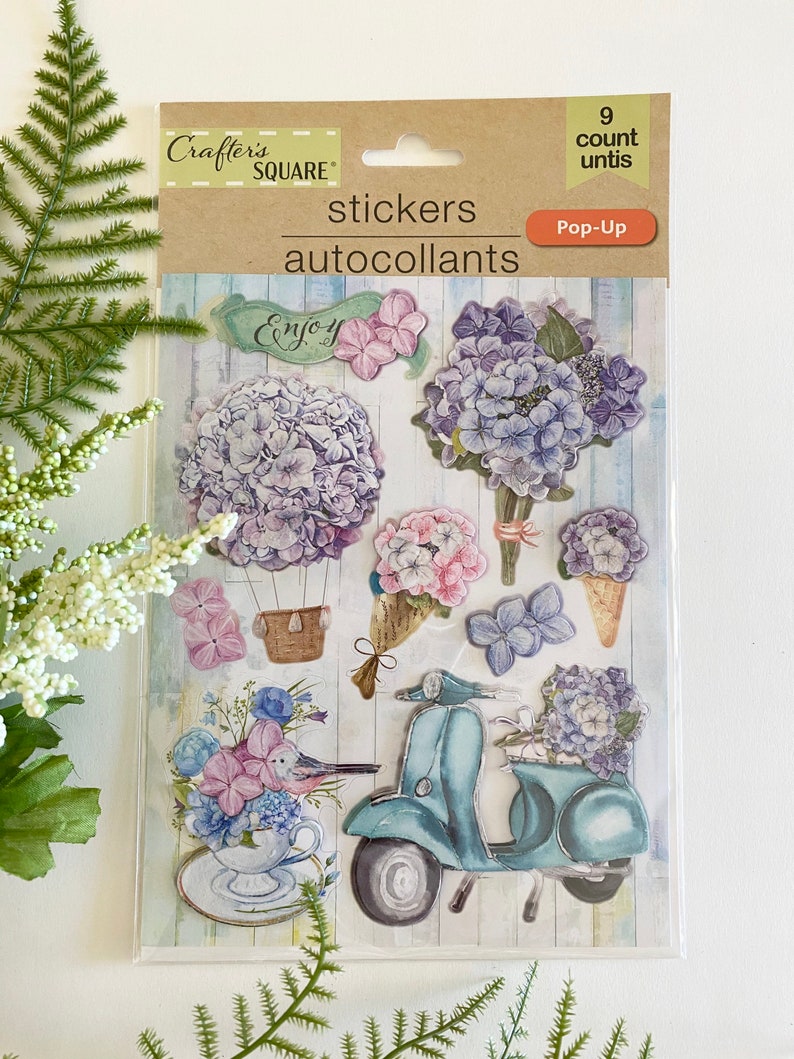 May include: A package of Crafter's Square pop-up stickers featuring floral designs, including hydrangeas, a scooter, and a teacup with a bird. The package includes the text "stickers" and "autocollants".