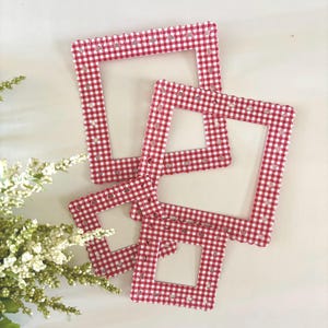May include: Four red and white gingham patterned picture frames with heart-shaped cutouts. The frames are arranged on a white surface, with a sprig of white flowers in the lower left corner.