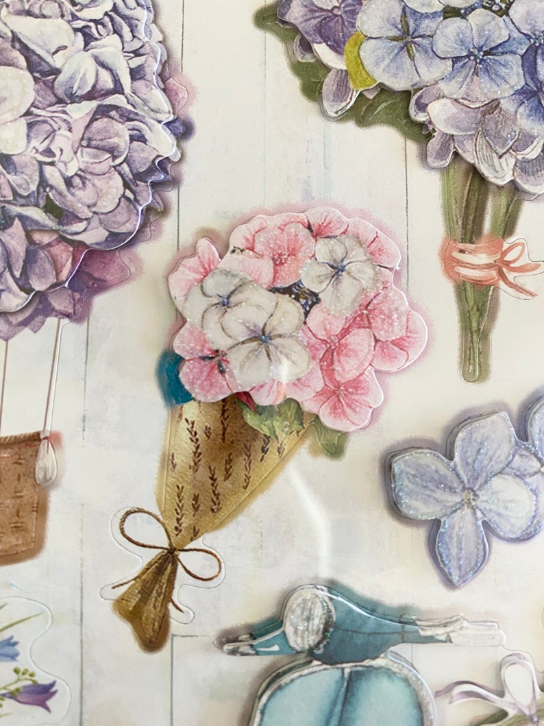 May include: A collection of floral stickers featuring various hydrangea bouquets in shades of purple, pink, and white. Some bouquets are tied with ribbons, and one is in a decorative cone. A blue scooter sticker is also visible.