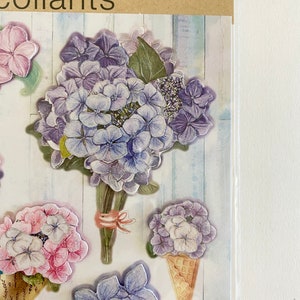 May include: Pop-up stickers featuring various hydrangea flower designs in shades of purple, pink, and white. The stickers include a bouquet, individual blooms, and a flower in an ice cream cone shape. The background has a light blue and white wood grain pattern.