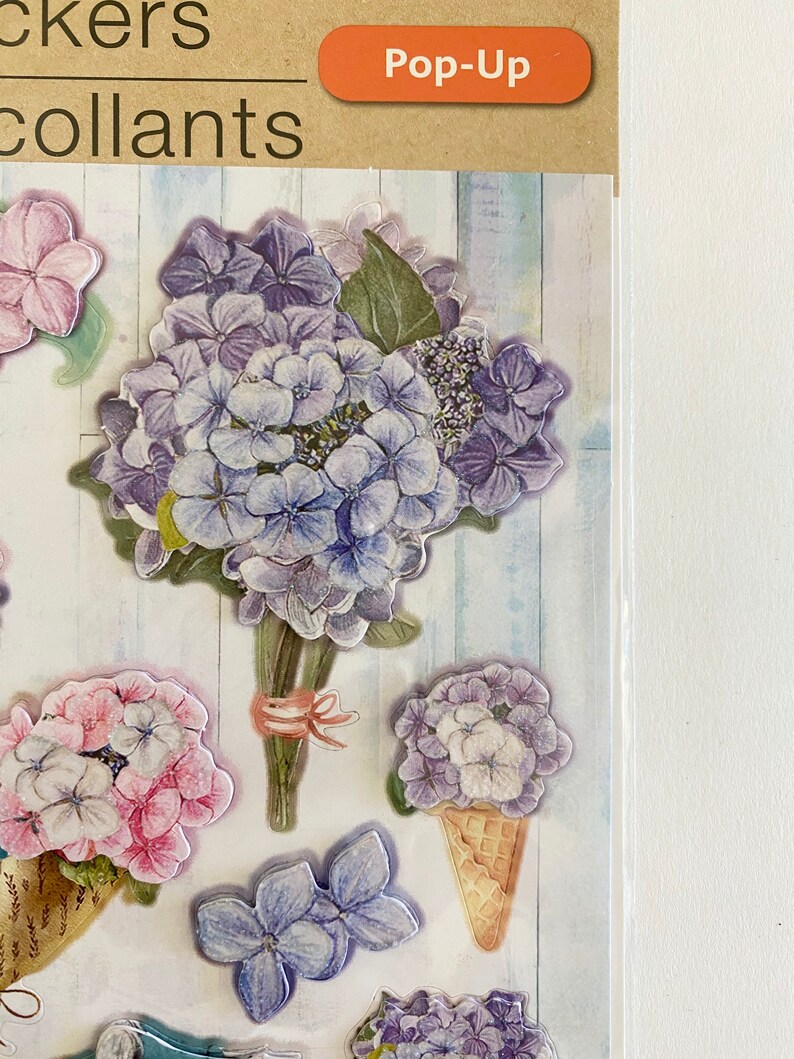 May include: Pop-up stickers featuring various hydrangea flower designs in shades of purple, pink, and white. The stickers include a bouquet, individual blooms, and a flower in an ice cream cone shape. The background has a light blue and white wood grain pattern.