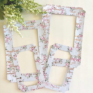 May include: A collection of decorative paper frames in various sizes, featuring a floral pattern with pink roses and green leaves on a light blue background. The frames have a vintage aesthetic with gold script accents.