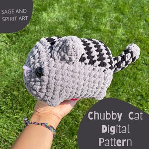 May include: A grey and black crocheted cat plush toy with a pink nose and large black eyes. The toy is being held in a hand with a rainbow bracelet. The text "Chubby Cat Digital Pattern" is visible in the image.