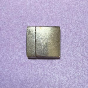 May include: A silver-colored rectangular magnetic clasp. The clasp is composed of two connected pieces, with a slightly textured surface. The background is a light purple color.