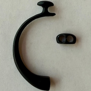 May include: Black earbud accessory set, including a curved ear hook and a small, oval-shaped piece with two holes. The ear hook has a rounded top and a curved design. The pieces are made of a matte black material.