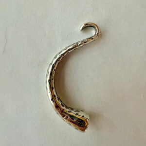 May include: A silver-colored, curved hook with a textured surface. The hook has a hammered appearance and a rounded end. The design is reminiscent of a horn or a stylized shape, suitable for jewelry or decorative purposes.