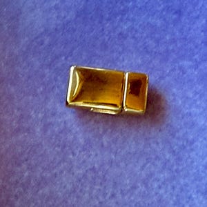 May include: A gold-coloured rectangular clasp with a smaller rectangular piece attached. The clasp is on a purple background. The clasp appears to be made of metal and has a shiny finish. The clasp is likely used for jewellery or other accessories.