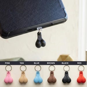 May include: A black phone charm shaped like a pair of testicles, attached to a phone case. Below, a row of keychains in pink, tan, blue, brown, black, and red. Each keychain has a similar testicle shape.