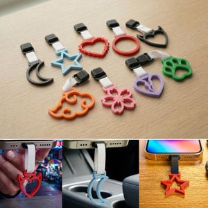 May include: A collection of phone charms in various shapes and colors, including a crescent moon, star, heart, ring, and paw print. Each charm has a black clip and a white strap for attaching to a phone.