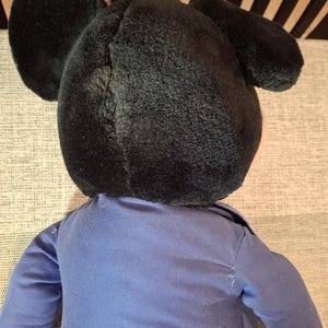 May include: A plush toy featuring a black head with large, round ears. The toy is wearing a blue shirt with long sleeves. The toy's design is reminiscent of a popular cartoon character.