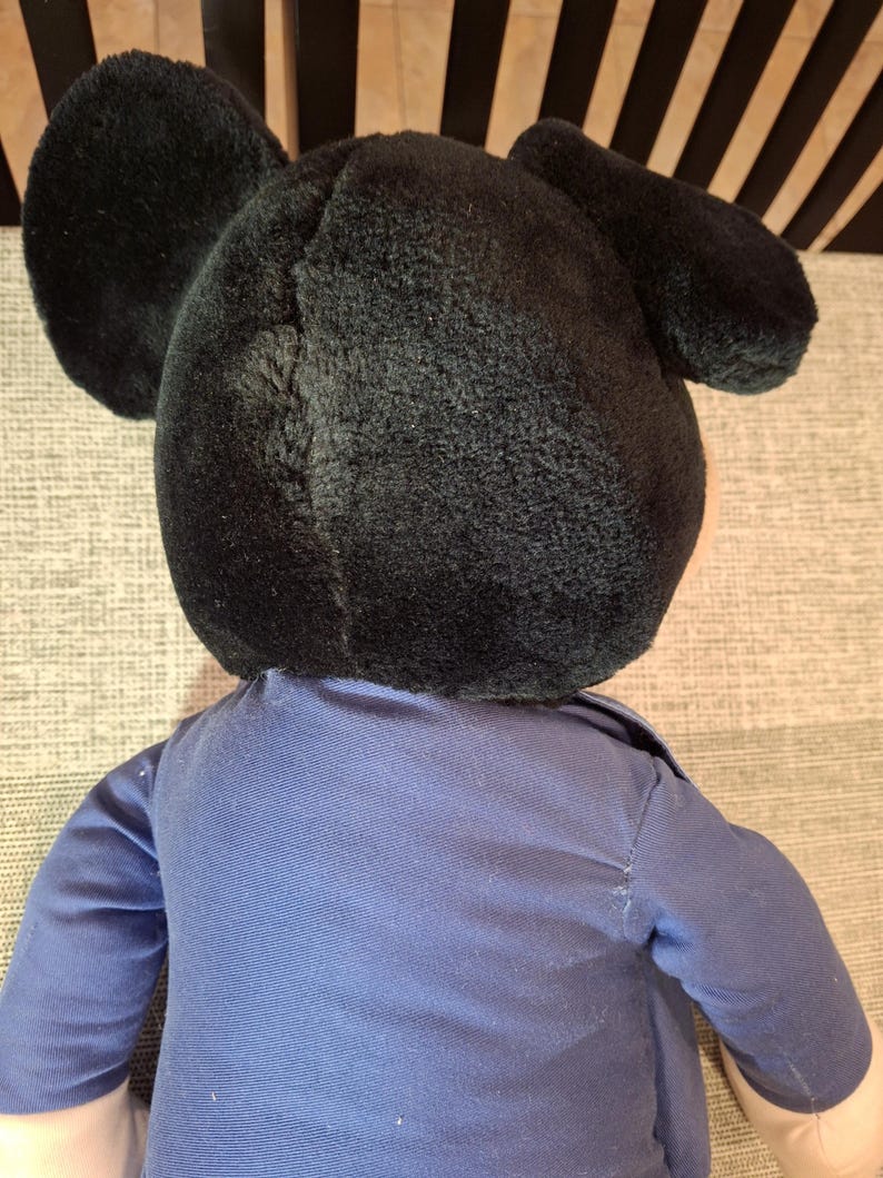 May include: A plush toy featuring a black head with large, round ears. The toy is wearing a blue shirt with long sleeves. The toy's design is reminiscent of a popular cartoon character.