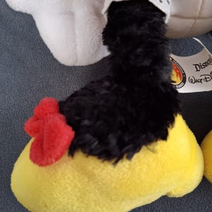 May include: Close-up of a plush toy foot. The foot is yellow with a red bow and black fur. A white lace trim is visible. The toy is a Disney Parks original.