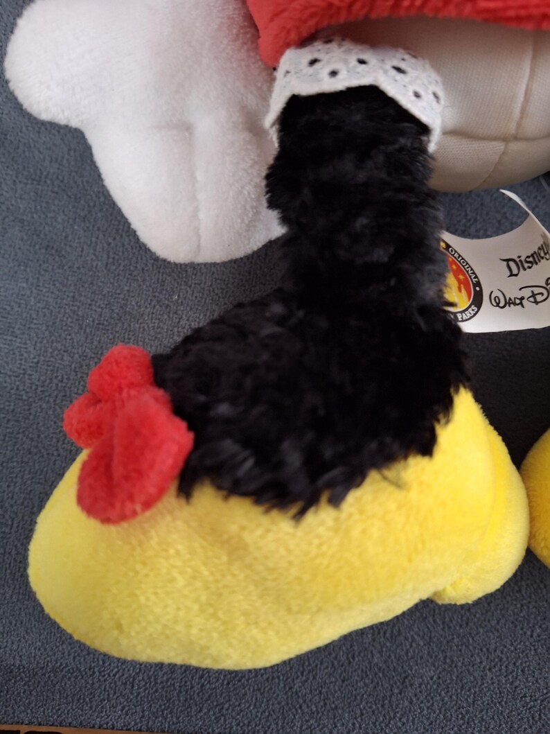 May include: Close-up of a plush toy foot. The foot is yellow with a red bow and black fur. A white lace trim is visible. The toy is a Disney Parks original.