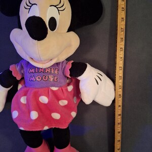 May include: A plush Minnie Mouse doll with a pink polka-dot skirt and bow. The doll is wearing a purple shirt with the words "MINNIE MOUSE" and red shoes. The doll is approximately 18 inches tall.