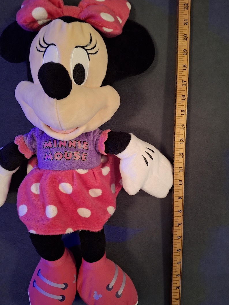 May include: A plush Minnie Mouse doll with a pink polka-dot skirt and bow. The doll is wearing a purple shirt with the words "MINNIE MOUSE" and red shoes. The doll is approximately 18 inches tall.