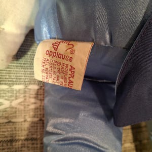 May include: A close-up of a plush toy with a blue satin fabric and a white fabric. A tag from "applause" is visible, with text including "WOODLAND K" and "MADE IN ME".