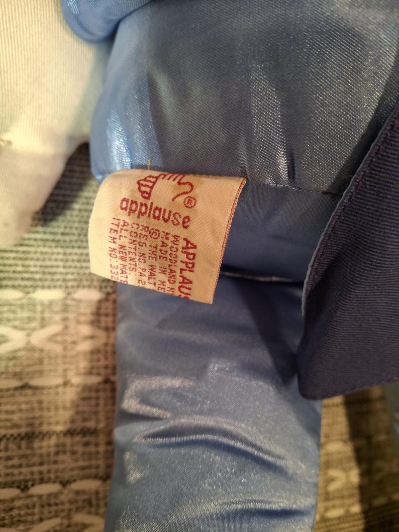 May include: A close-up of a plush toy with a blue satin fabric and a white fabric. A tag from "applause" is visible, with text including "WOODLAND K" and "MADE IN ME".