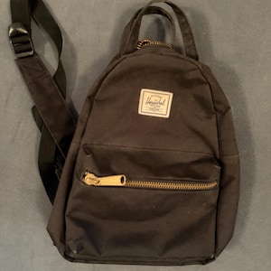 May include: A black Herschel backpack featuring a top handle, adjustable black shoulder straps, and a front zippered pocket with a gold-toned metal zipper. A white rectangular brand label with black text is stitched onto the upper front section.