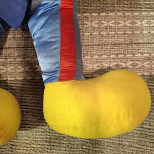 May include: Close-up of a plush toy's leg and foot. The leg is blue satin with a red stripe down the side. The foot is a large, rounded yellow shape. The toy is resting on a patterned gray surface.