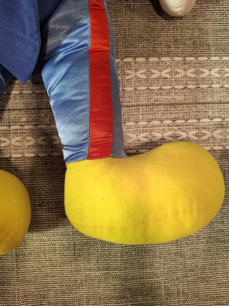 May include: Close-up of a plush toy's leg and foot. The leg is blue satin with a red stripe down the side. The foot is a large, rounded yellow shape. The toy is resting on a patterned gray surface.