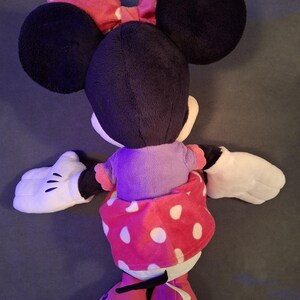 May include: A plush Minnie Mouse doll, viewed from the back, features large black ears and a red polka-dot bow. She wears a purple top and a red polka-dot skirt. White gloves and red shoes complete the look.