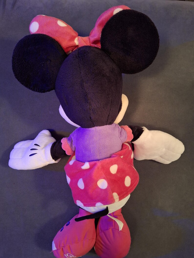 May include: A plush Minnie Mouse doll, viewed from the back, features large black ears and a red polka-dot bow. She wears a purple top and a red polka-dot skirt. White gloves and red shoes complete the look.