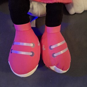 May include: Close-up of a plush toy's legs and feet. The feet are bright pink with white soles and silver accents. The legs are black, and the toy is wearing a red polka-dot skirt.
